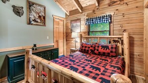 A wooden cabin bedroom with a bed, a dresser, and a window with a checkered curtain.