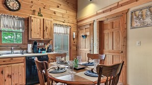 A wooden interior with a dining table set for four, wooden cabinets, a clock, and a window with blinds.