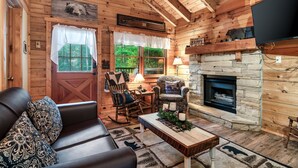 A cozy log cabin interior with a stone fireplace, wooden walls, a leather sofa, and a rocking chair.