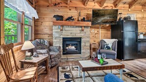 A cozy cabin interior with a stone fireplace, wooden walls, a television, and a dining area.