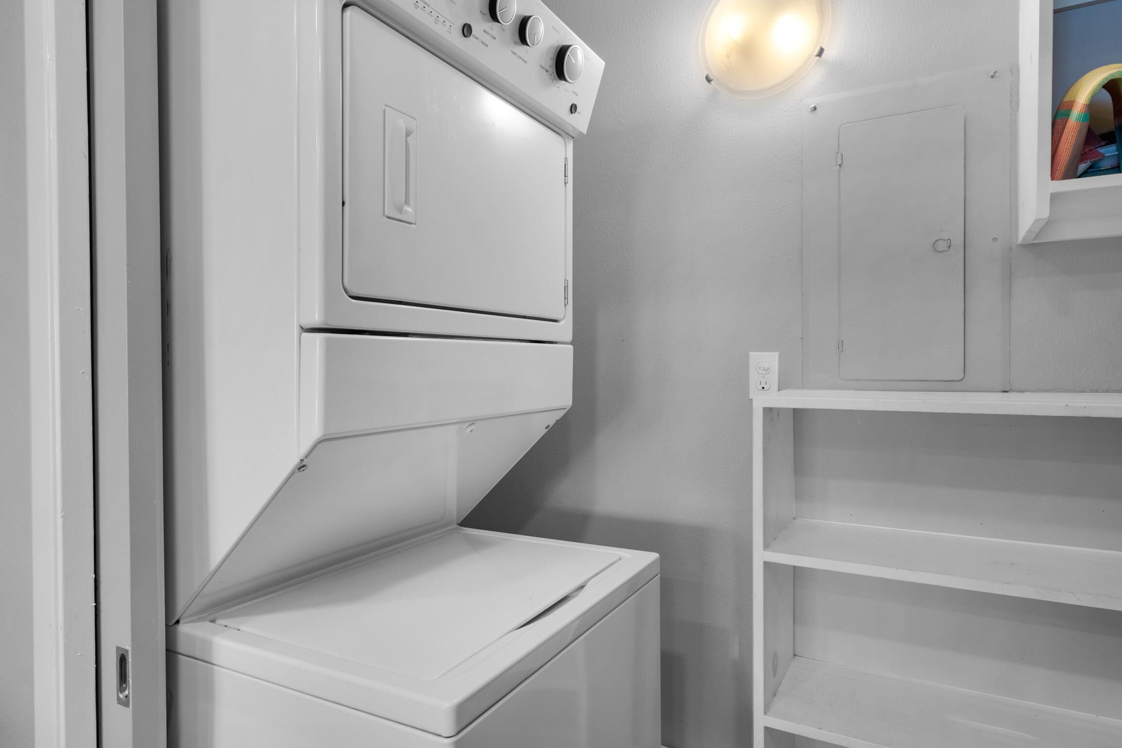 A laundry room with a stacked washer and dryer, a shelf with cleaning supplies, and a wall-mounted light fixture.