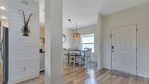Una cocina moderna con armarios blancos, un comedor con mesa y sillas de madera, y un pasillo con una puerta blanca y una alfombra.
