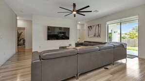 Una sala de estar moderna con un sofá seccional, un ventilador de techo, un televisor de pantalla plana y vistas al exterior.