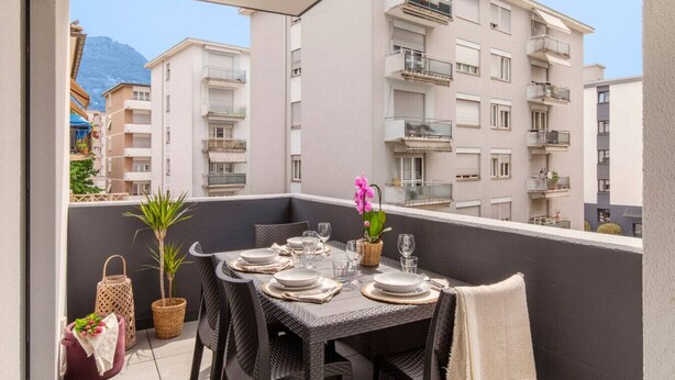 Un balcón con una mesa de comedor dispuesta para cuatro personas, sillas de mimbre, una pequeña planta en maceta y una cesta de mimbre.