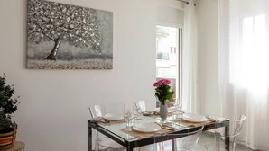 Comedor con mesa de vidrio, sillas transparentes, un florero con flores y un cuadro en la pared.