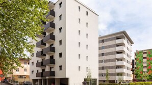 Un edificio residencial moderno de varios pisos con balcones, un área de estacionamiento techada y un edificio vecino con un mural colorido.