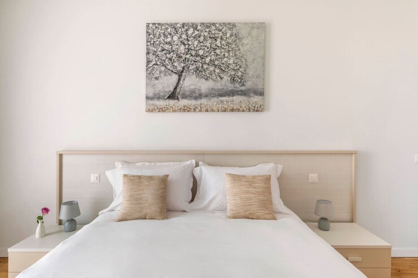 A bedroom with a bed, two bedside tables, a nightstand, a lamp, and a wall-mounted painting of a tree.