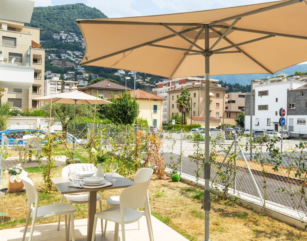 Une terrasse extérieure avec une table et des chaises, un grand parasol et une vue sur un quartier résidentiel avec des immeubles et une route.