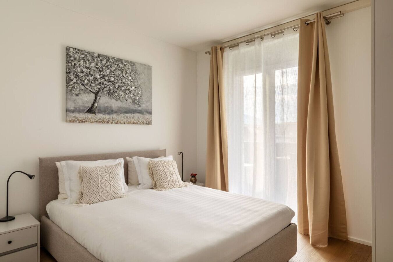 A neatly made bed with a white duvet and two decorative pillows, a bedside table with a lamp, and a large window with beige curtains.
