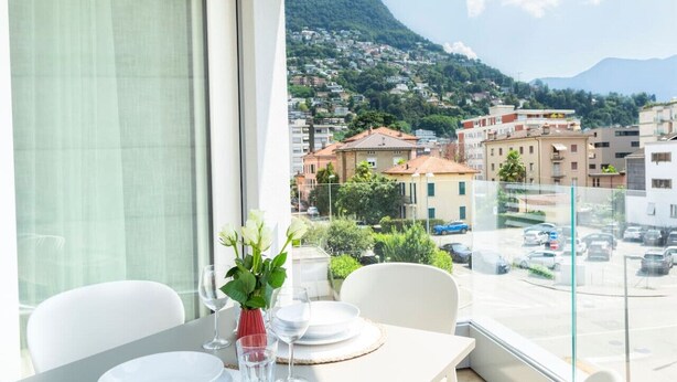 A dining area with a table set for two, a vase with flowers, and a view of a hillside town. - Elegant new Apt with AC, private balcony & garage (Viganello)