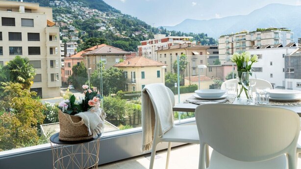 A balcony with a table set for dining, overlooking a cityscape with buildings and a mountain. - Elegant new Apt with AC, private balcony & garage (Viganello)
