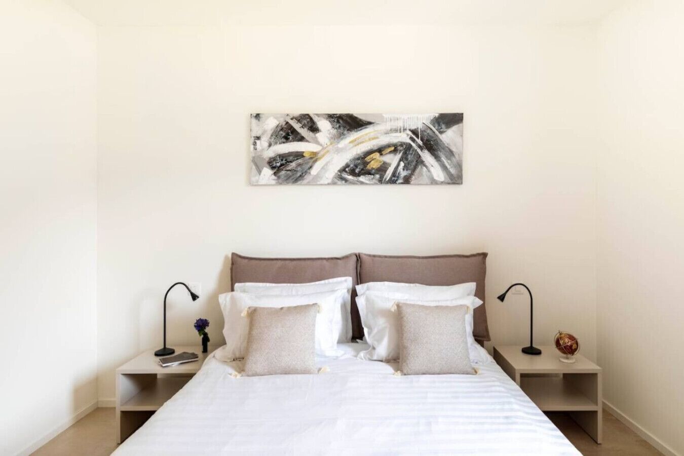 A bedroom with a bed, two bedside tables, a nightstand, a wall-mounted abstract painting, and a small decorative globe.