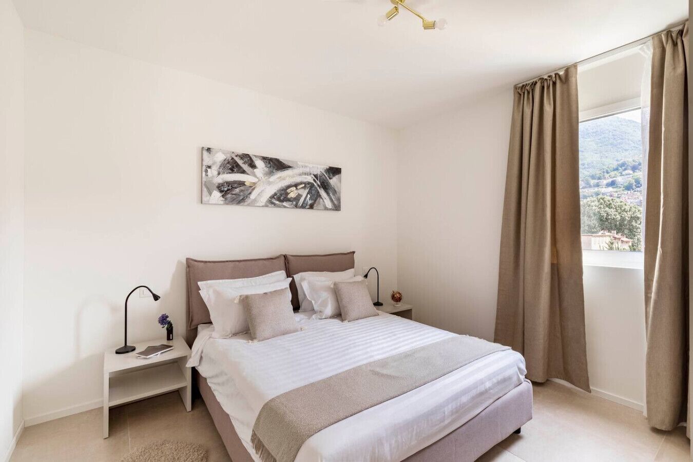 Ein Schlafzimmer mit einem großen Bett, zwei Nachttischen, einer Stehlampe, einer Deckenleuchte, einem Fenster mit Vorhängen und Blick ins Freie.