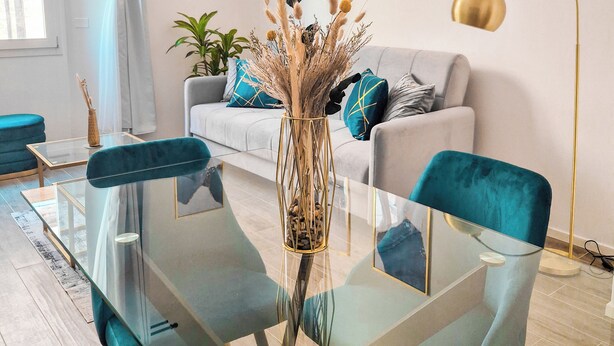 A modern living room with a glass coffee table, teal chairs, a white sofa with blue cushions, and a floor lamp. - Ginevra Suite (Bologna)