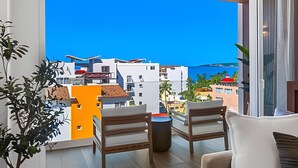 A balcony with two chairs and a small table, overlooking a coastal town with white buildings and a clear blue sky. - 1 BR Golden Zone · Luxury Beach and Infinity Pool (Bucerías)