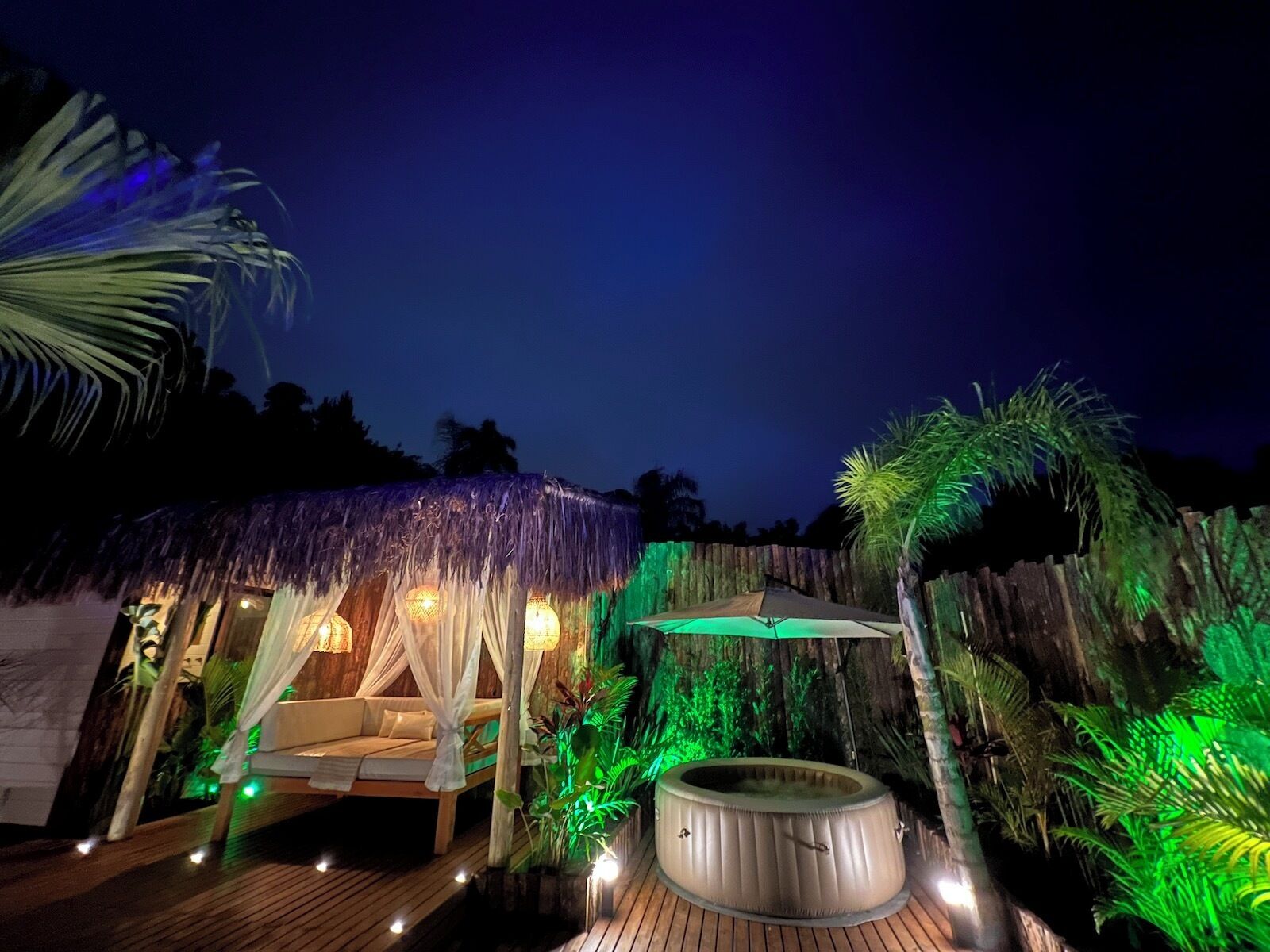 Una cabaña con techo de paja, con cama y bañera de hidromasaje, rodeada de plantas tropicales y una terraza de madera.