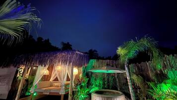 Una cabaña con techo de paja, con cama y bañera de hidromasaje, rodeada de plantas tropicales y una terraza de madera.