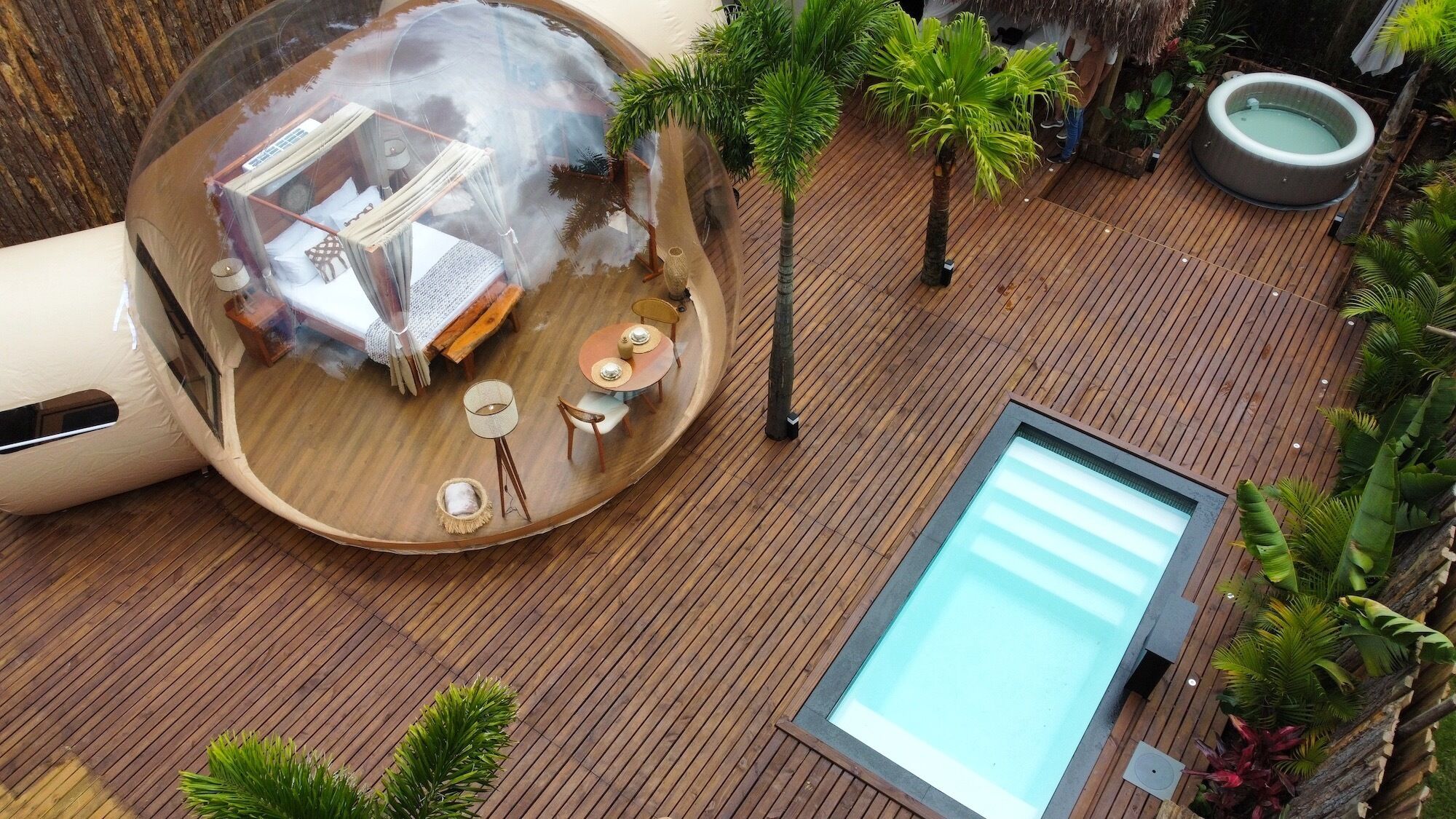 A circular wooden deck with a glass-enclosed room containing a bed, a table, and chairs. There is a pool and a hot tub on the deck.