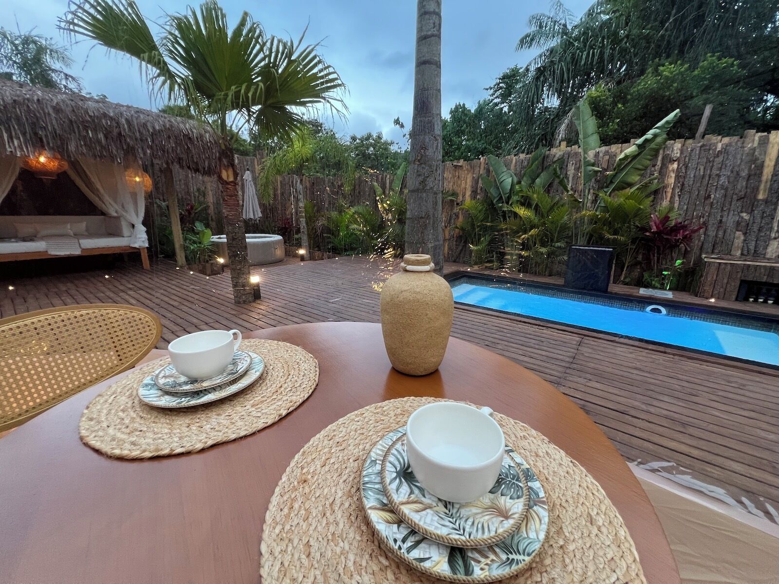 Terraza de madera con piscina, asientos con techo de paja y una mesa de comedor con dos tazas y platillos blancos.