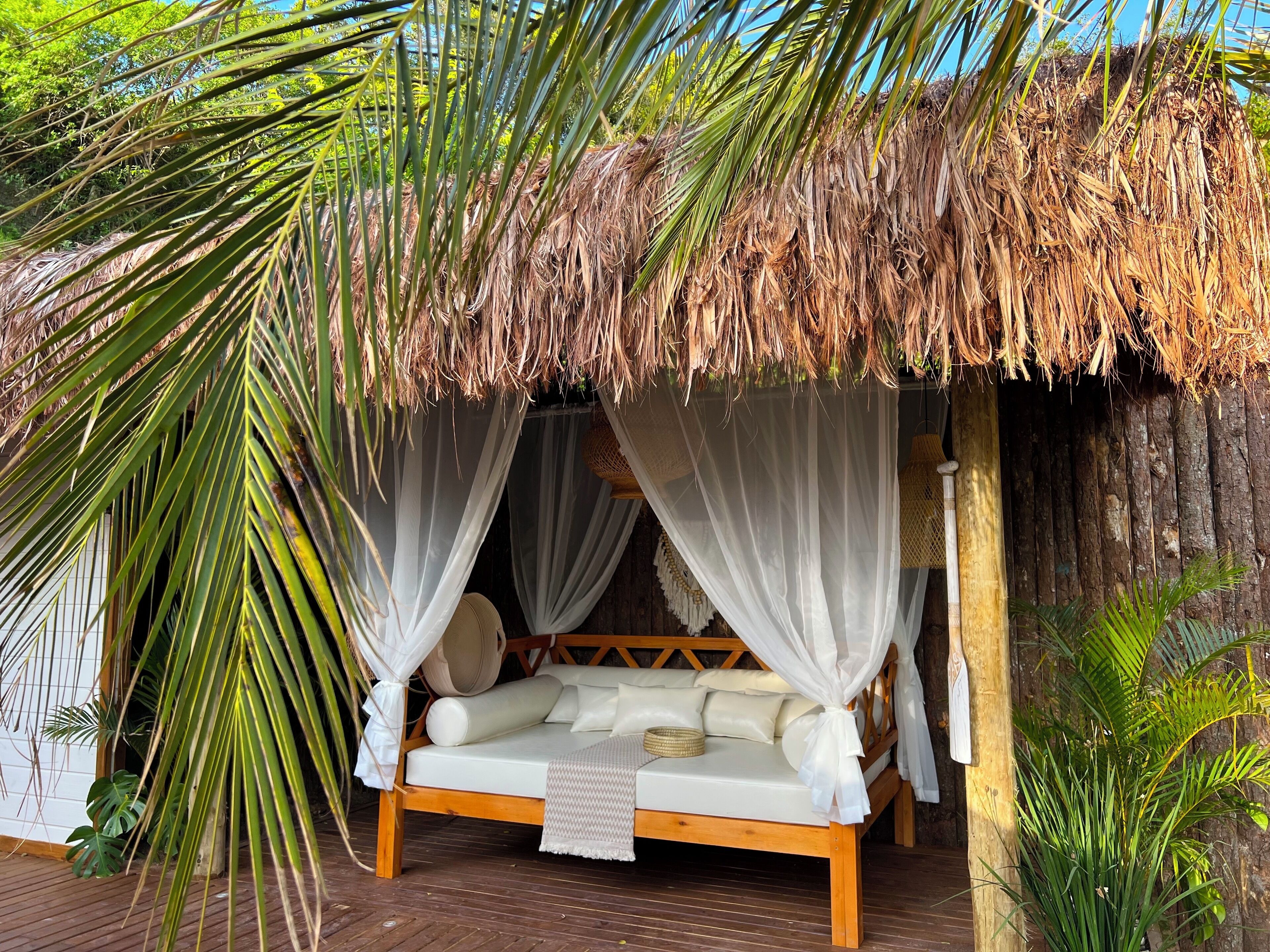 A thatched-roof outdoor bedroom with a wooden bed and white canopy.