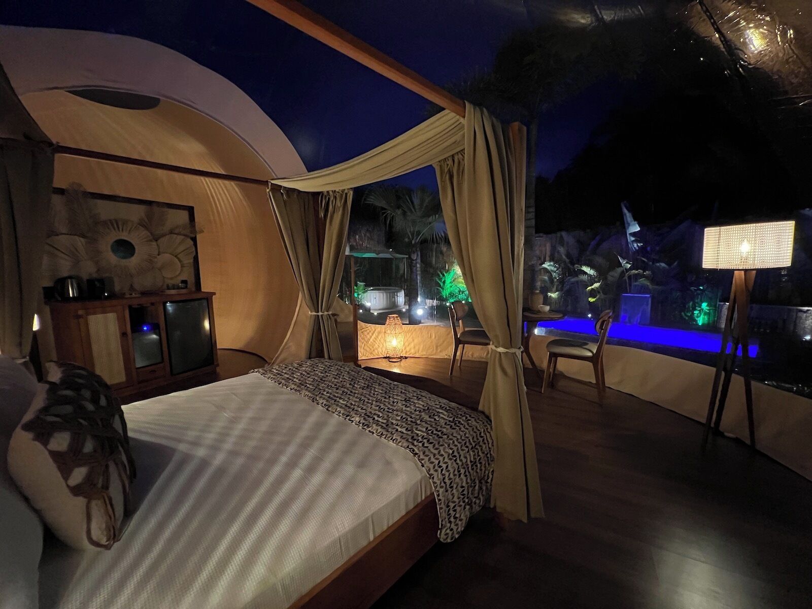 A tented room with a bed, a small cabinet, and a view of an outdoor pool area with lights.