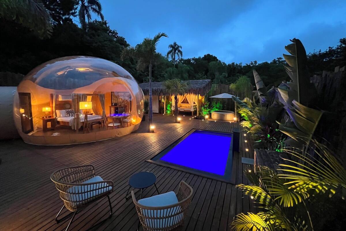 A spherical glass yurt with a wooden deck, a pool, and wicker furniture.