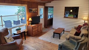 A cozy cabin interior with a wooden floor, a sofa, a TV on a wooden cabinet, and a view of the lake through large windows. - Rustic Retreat on Mississagua Lake in Beautiful Trent Lakes (Trent Lakes)