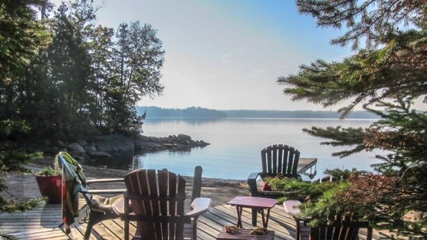 A wooden deck with Adirondack chairs and a table overlooking a calm lake, surrounded by trees. - Rustic Retreat on Mississagua Lake in Beautiful Trent Lakes (Trent Lakes)