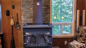A wood-burning stove with the brand name 'KANATA' in a rustic room with a stone wall and a window with a view of trees. - Rustic Retreat on Mississagua Lake in Beautiful Trent Lakes (Trent Lakes)