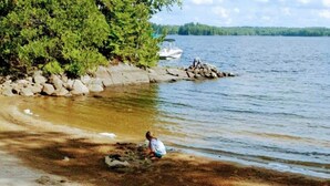 A sandy beach with a person near the water's edge, a boat in the background, and a forested shoreline. - Rustic Retreat on Mississagua Lake in Beautiful Trent Lakes (Trent Lakes)