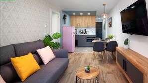 A compact living space with a grey sofa, a small round wooden table, a pink refrigerator, and a kitchen area with wooden cabinets and a TV mounted on the wall.