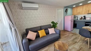 A modern living room with a grey sofa, a small round table, and a kitchen area with wooden cabinets and a pink refrigerator.