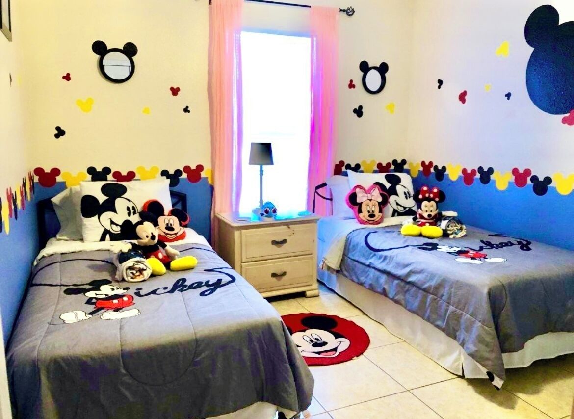 Une chambre d’enfant avec deux lits, chaque couvre-lit représentant Mickey Mouse, une commode, une lampe et des décorations murales comprenant des silhouettes de Mickey Mouse et des cœurs.