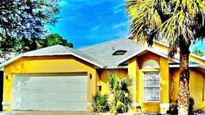 A yellow single-story house with a garage, a small porch, and a palm tree. - Ref 319. Modern lake view pool home. Very close to Disney (Kissimmee)