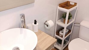 Casa de banho com lavatório branco, sanita e um armário branco com cestos e rolo de papel higiénico.