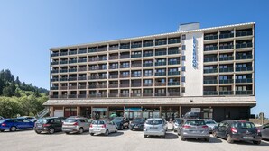 Een hotel met meerdere verdiepingen, een centraal entree, diverse balkons en een parkeerplaats met verschillende auto's.