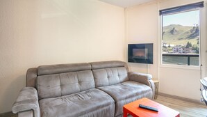 Een woonkamer met een grijze bank, een kleine rode tafel en een aan de muur bevestigde televisie.