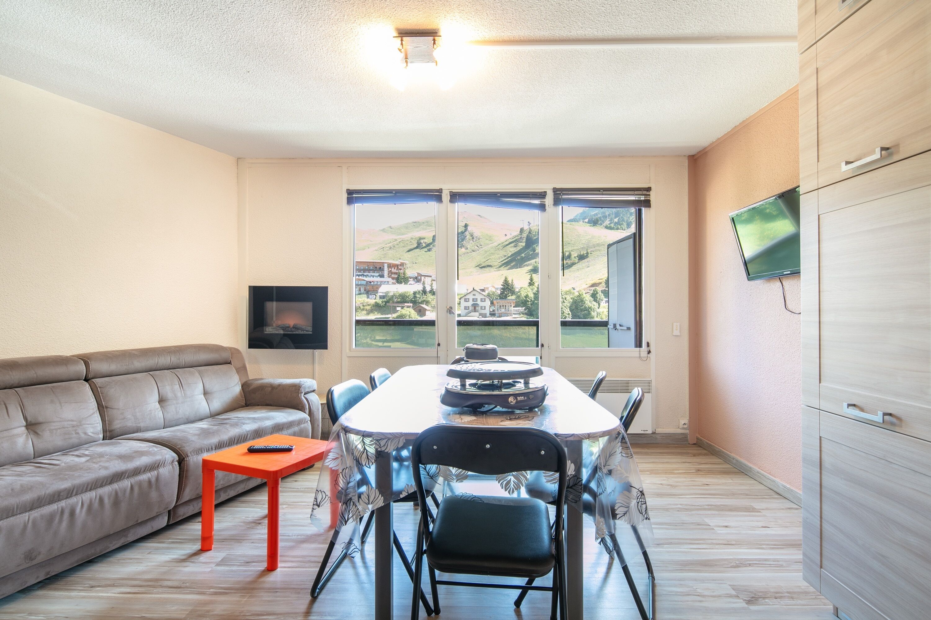 Un salon moderne comprenant un canapé, une table à manger avec des chaises, un téléviseur fixé au mur et de grandes fenêtres offrant une vue sur les collines.