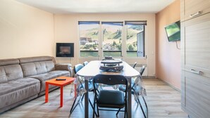 Een moderne woonkamer met een bank, een eettafel met stoelen, een aan de muur bevestigde televisie en grote ramen met uitzicht op de heuvels.