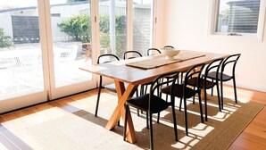Un área de comedor con mesa de madera y sillas negras, ventanas amplias con vistas a un patio trasero y una alfombra tejida en el suelo.