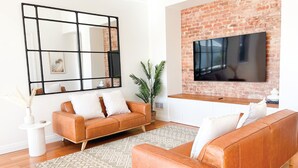 Un salón con pared de ladrillo, un televisor de pantalla plana grande, un sofá de cuero marrón y una alfombra estampada.