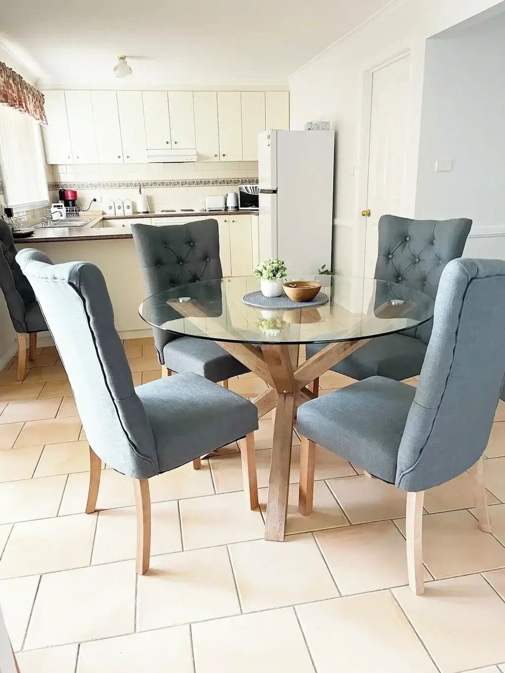 A modern kitchen with white cabinets, a white refrigerator, and a dining area with a glass table and four grey upholstered chairs.