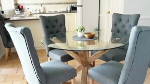A modern kitchen with white cabinets, a white refrigerator, and a dining area with a glass table and four grey upholstered chairs.