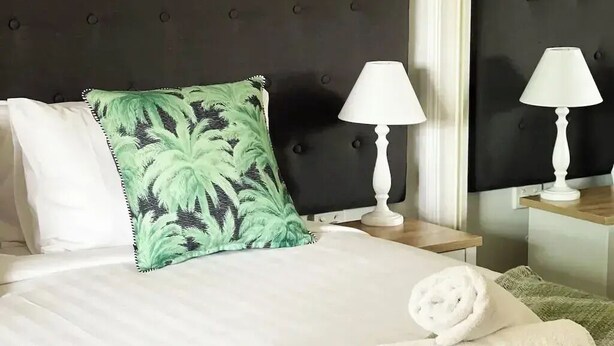 A neatly made bed with white linens, a green patterned pillow, and a rolled white towel on a green herringbone throw.
