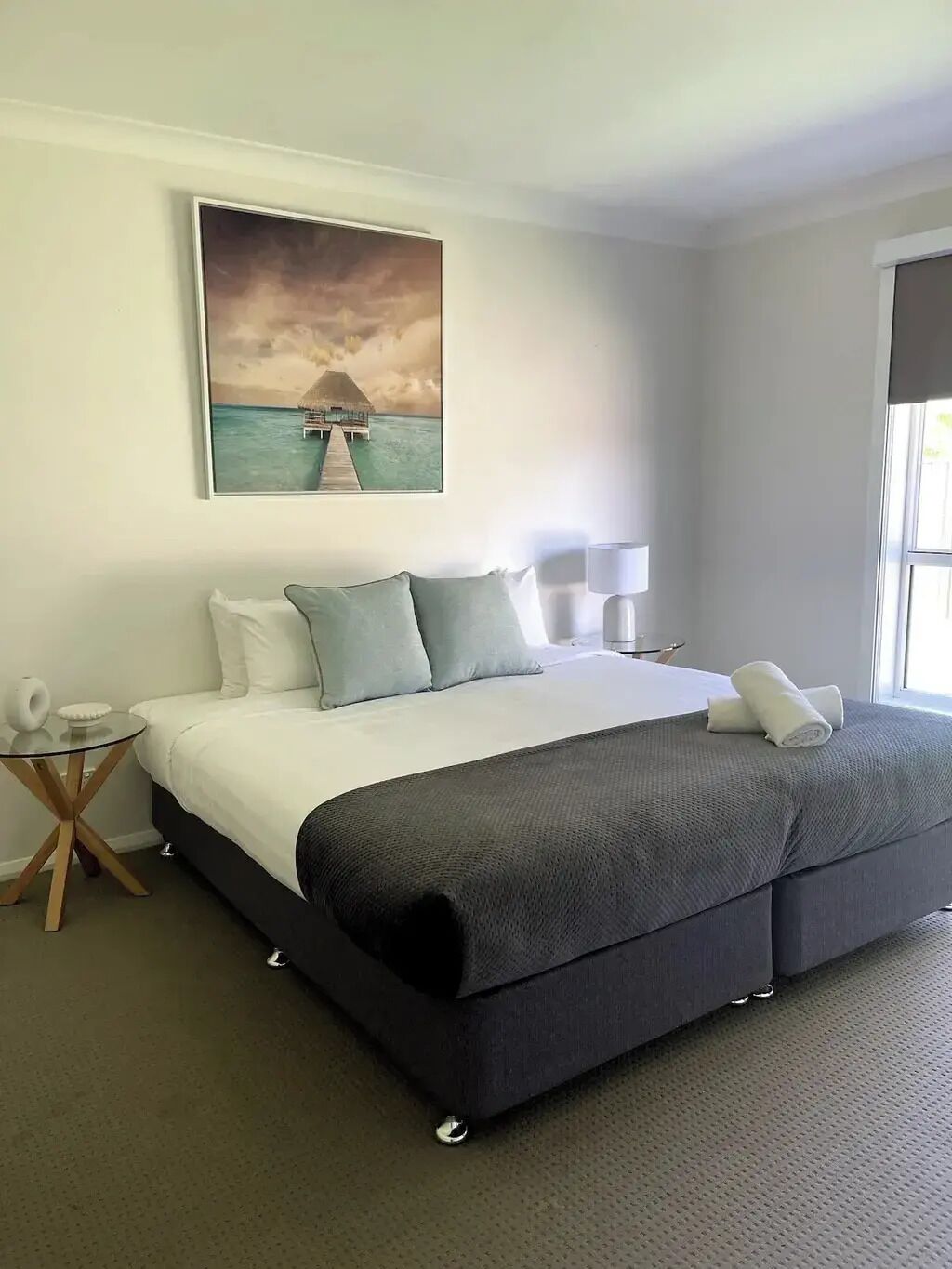 A hotel room with a large bed, a painting of a tropical scene, a small side table, and a lamp.