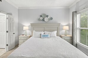 A bedroom with a large bed, two bedside tables, a nightstand, a wall clock, and a window with blinds.
