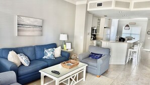 Un salon avec un canapé bleu, un fauteuil à rayures, une table basse en verre et un coin cuisine visible à travers une porte ouverte.