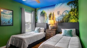 A room with two beds, a window with curtains, a wall mural, and a nightstand with a lamp.