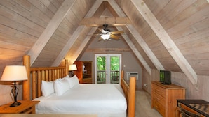 A cozy attic bedroom with a wooden ceiling, a bed with white linens, a desk with a computer, and a window with a view of greenery. - Smoky Mtn Escape | Indoor Pool, Arcade, Theater & Firepit Nights (Gatlinburg)