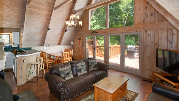 A cozy cabin interior with a dining area, a sofa with decorative pillows, a TV, and large windows offering a view of the outdoors. - Smoky Mtn Escape | Indoor Pool, Arcade, Theater & Firepit Nights (Gatlinburg)